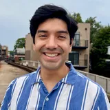 Érick smiling outdoors in a blue and white striped shirt, with an elevated train track and apartment buildings behind him.